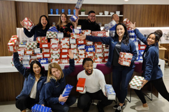 A diverse group of adults who work at one of Shoes That Fit's corporate sponsors, posing with enthusiasm with approximately 100 boxes of shoes they've collected to donate