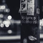 a bunch of stickers on a pole on the street. the most prominent one reading "big data is watching you". background is a cityscape