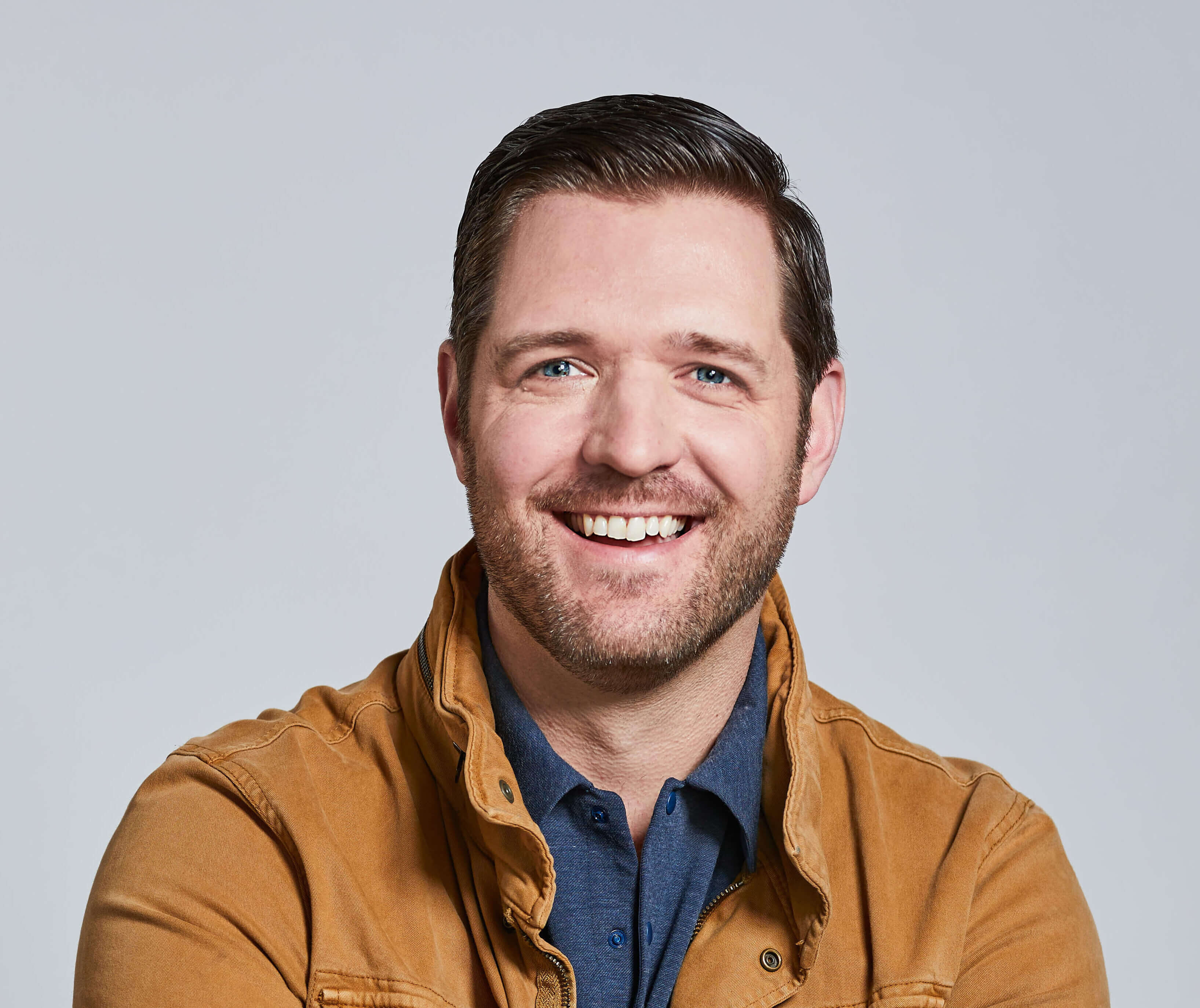 Headshot of smiling man with beard. Wearing a camel colored utility jacket and blue button up collard shirt underneath.