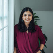 Smiling photo of Meena Das, who has long dark hair and is wearing a burgundy wrap style top and black pants. She is leaning against a wall near a window and is bathed in natural light.