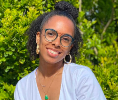 In this headshot, Devyn smiles and is wearing hoop earrings and a gold necklace with a green charm.