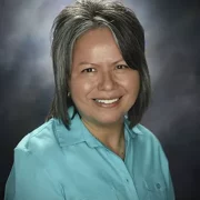 Sharon Herrera posing for headshot in front of grey background. She has early length hair with brown eyes.