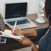A person of color works from home wearing white headphones and writing in a notebook