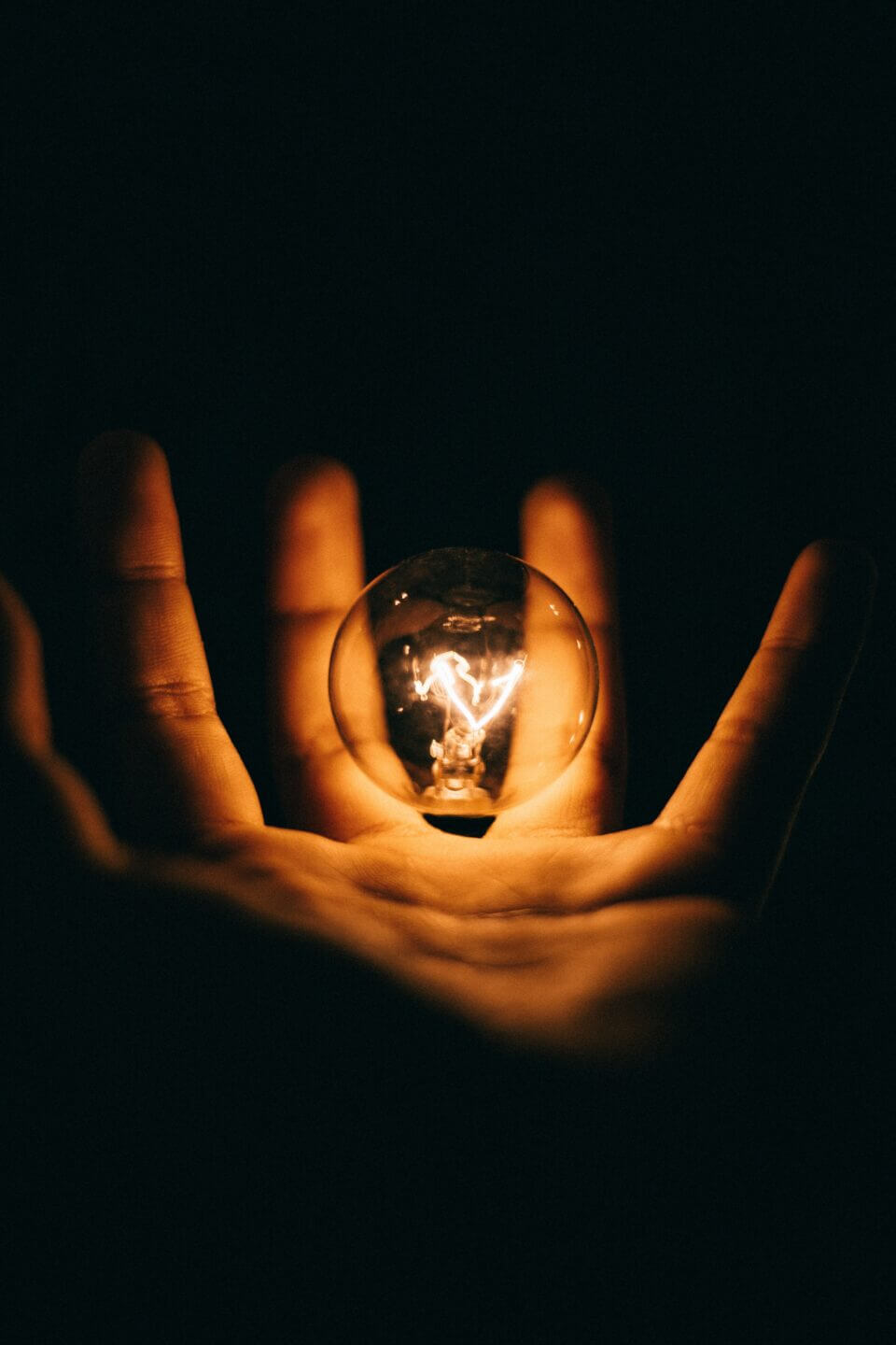 A close up of a person's hand. It is dark and the light bulb floating just above their open palm provides the only illumination.
